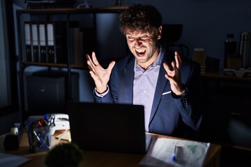 Hispanic young man working at the office at night crazy and mad shouting and yelling with aggressive expression and arms raised. frustration concept.