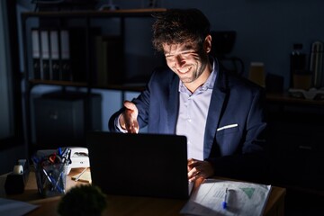 Hispanic young man working at the office at night smiling friendly offering handshake as greeting and welcoming. successful business.