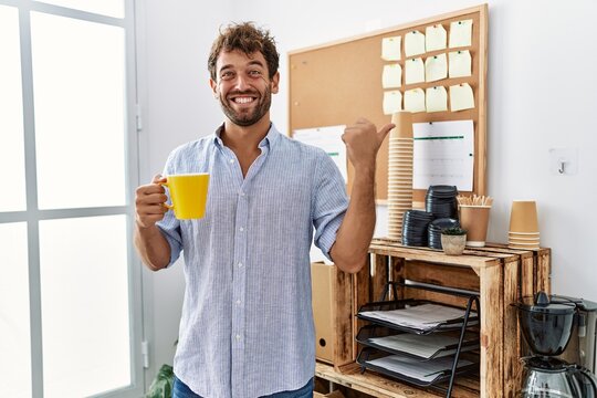 Young handsome man drinking a cup coffee at the office pointing thumb up to the side smiling happy with open mouth