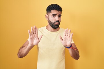 Hispanic man with beard standing over yellow background moving away hands palms showing refusal and...