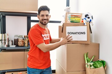 Young man with beard wearing volunteer t shirt at donations stand looking positive and happy...