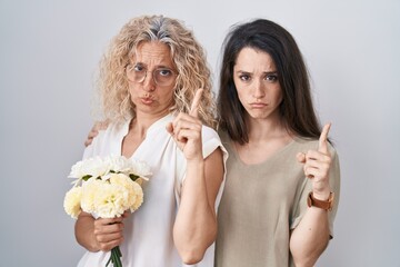 Mother and daughter holding bouquet of white flowers pointing up looking sad and upset, indicating...