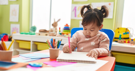 Adorable chinese girl preschool student sitting on table drawing on notebook at kindergarten