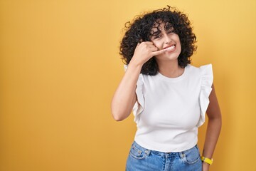 Young middle east woman standing over yellow background smiling doing phone gesture with hand and fingers like talking on the telephone. communicating concepts.