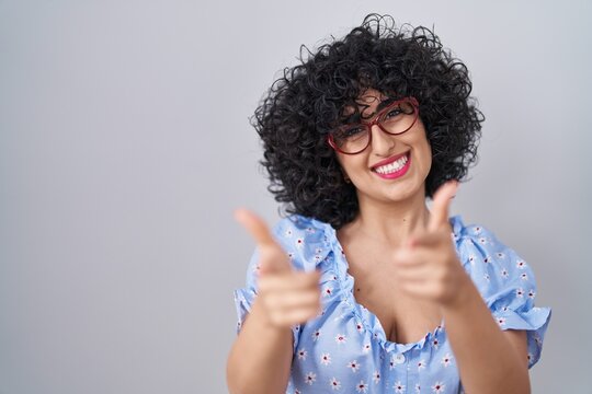 Young brunette woman with curly hair wearing glasses over isolated background pointing fingers to camera with happy and funny face. good energy and vibes.