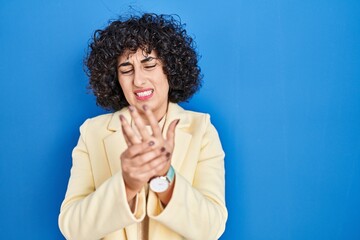 Young brunette woman with curly hair standing over blue background suffering pain on hands and...