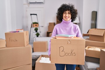 Young brunette woman with curly hair moving to a new home holding cardboard box winking looking at the camera with sexy expression, cheerful and happy face.