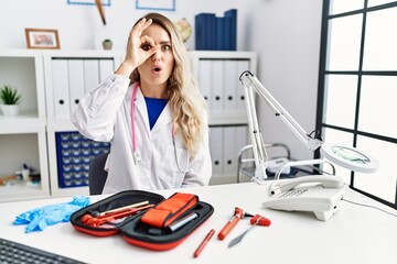 Young beautiful doctor woman with reflex hammer and medical instruments doing ok gesture shocked...