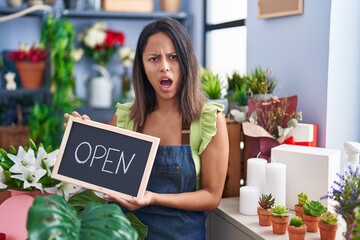 Hispanic young woman working at florist with open sign in shock face, looking skeptical and sarcastic, surprised with open mouth