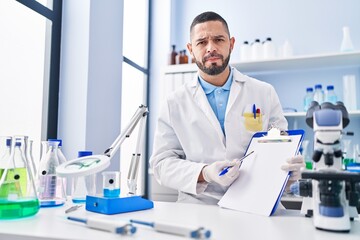 Hispanic man working at scientist laboratory holding blank clipboard skeptic and nervous, frowning upset because of problem. negative person.