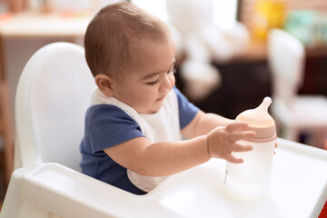 Adorable chinese toddler holding feeding bottle sitting on dinner chair baby at home
