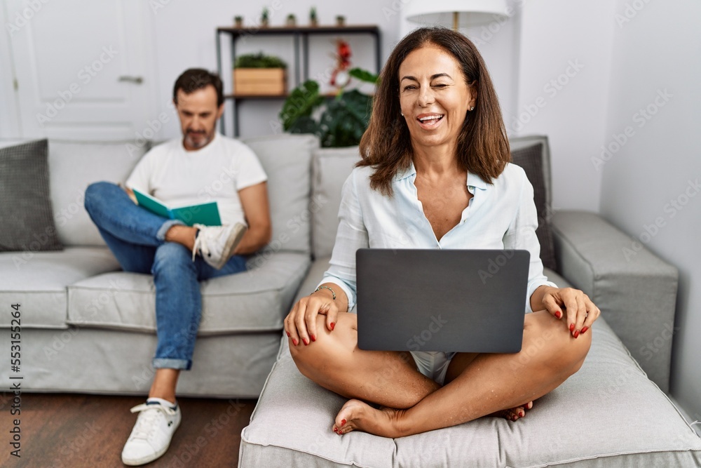 Sticker Hispanic middle age couple at home, woman using laptop winking looking at the camera with sexy expression, cheerful and happy face.