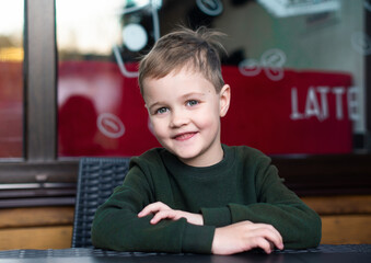 The boy sits at the table and looks straight ahead. The light-haired boy is 6 years old. He is wearing a green sweater and smiling. The photo is blurred