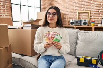 Young hispanic woman sitting on the sofa at new home holding money smiling looking to the side and...