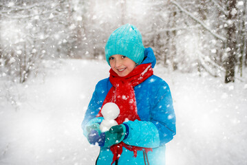 happy boy in winter bright clothes holds a snowball in his hand, walks through the snowy forest