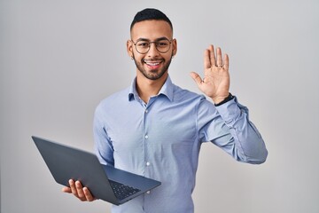 Young hispanic man working using computer laptop waiving saying hello happy and smiling, friendly...