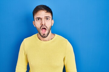 Obraz premium Hispanic man standing over blue background in shock face, looking skeptical and sarcastic, surprised with open mouth