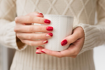 Woman's hands with red manicure and gold foil on the nails. Trendy autumn and winter nail design. Woman with a beautiful manicure holding a white knitted cup. The concept of cozy Christmas holidays. - obrazy, fototapety, plakaty