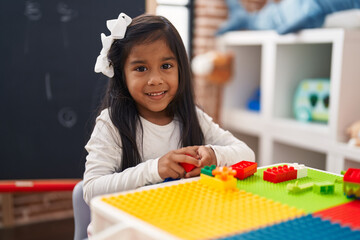 Adorable hispanic girl playing with construction blocks sitting on table at kindergarten