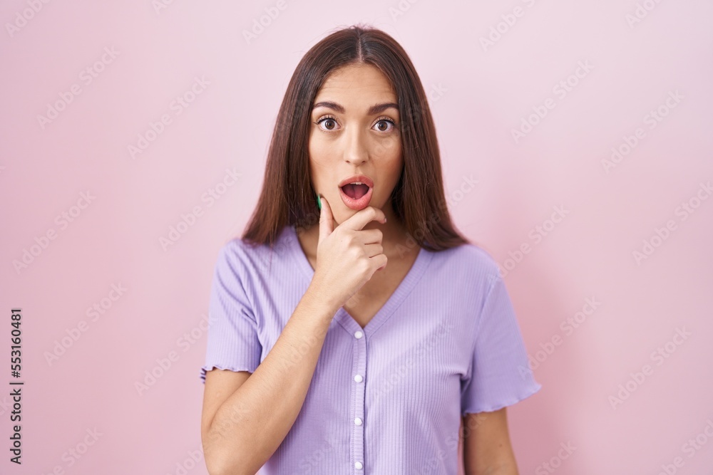 Wall mural Young hispanic woman with long hair standing over pink background looking fascinated with disbelief, surprise and amazed expression with hands on chin