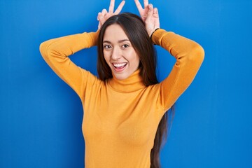 Fototapeta na wymiar Young brunette woman standing over blue background posing funny and crazy with fingers on head as bunny ears, smiling cheerful