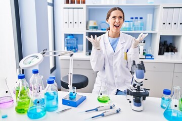 Young brunette woman working at scientist laboratory crazy and mad shouting and yelling with aggressive expression and arms raised. frustration concept.