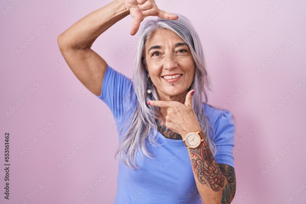 Poster Middle age woman with tattoos standing over pink background smiling making frame with hands and fingers with happy face. creativity and photography concept.