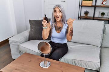 Middle age grey-haired woman using night serum sitting on the sofa pointing thumb up to the side smiling happy with open mouth