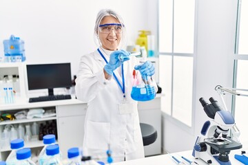 Middle age grey-haired woman wearing scientist uniform pouring liquid on test tube at laboratory