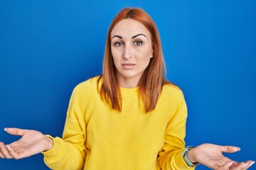 Young woman standing over blue background clueless and confused with open arms, no idea concept.