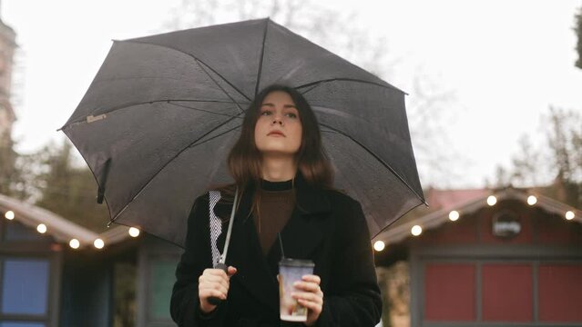 Young cute serious attractive woman girl goes for a walk on the city street in the rain, holds a black umbrella and drinks coffee