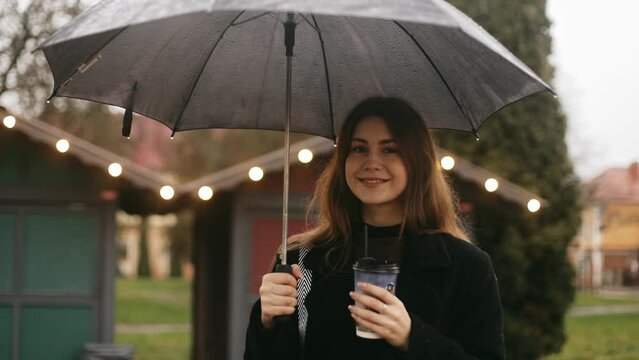 Young pretty cute smiling woman girl standing on a city street at a Christmas fair in the rain, holding a black umbrella and drinking coffee through a straw