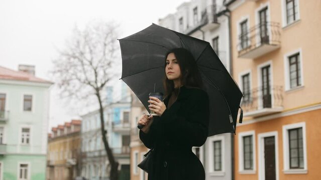 Portrait of a young cute smiling attractive woman girl standing alone on a city street in the rain, holding a black umbrella and drinking coffee through a straw, loves the rain
