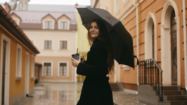 Rear view of young cute smiling attractive woman girl in coat walking alone on city street in rain, holding black umbrella and cup of coffee, turning to camera and smiling