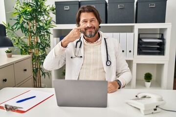 Handsome middle age doctor man working at the clinic pointing with hand finger to face and nose, smiling cheerful. beauty concept