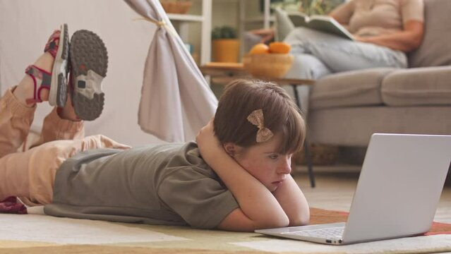 Girl with down syndrome lying on her stomach on floor in living room watching cartoon on laptop