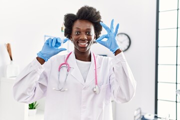 African doctor woman holding syringe at medical clinic doing ok sign with fingers, smiling friendly...