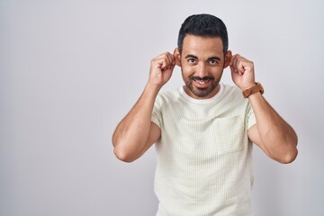 Hispanic man with beard standing over isolated background smiling pulling ears with fingers, funny...
