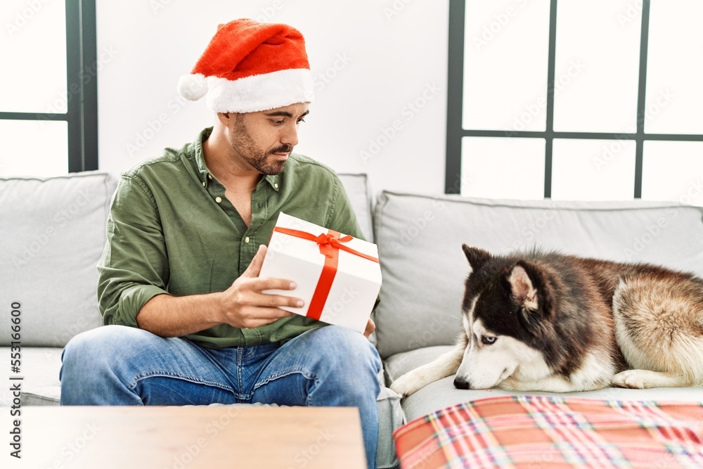 Poster Young hispanic man holding gift sitting on sofa with dog by christmas decor at home