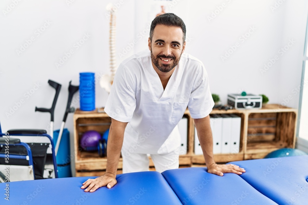 Sticker young hispanic man wearing physiotherapist uniform standing at clinic