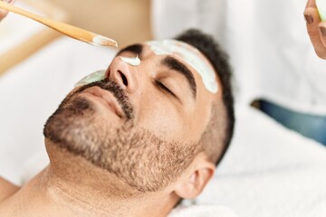 Young hispanic man having facial mask treatment at beauty center