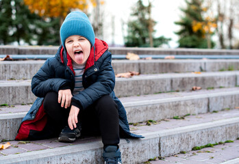 The boy is sitting on the stairs. He is 6 years old. He is wearing a jacket and a blue hat. A rowdy...