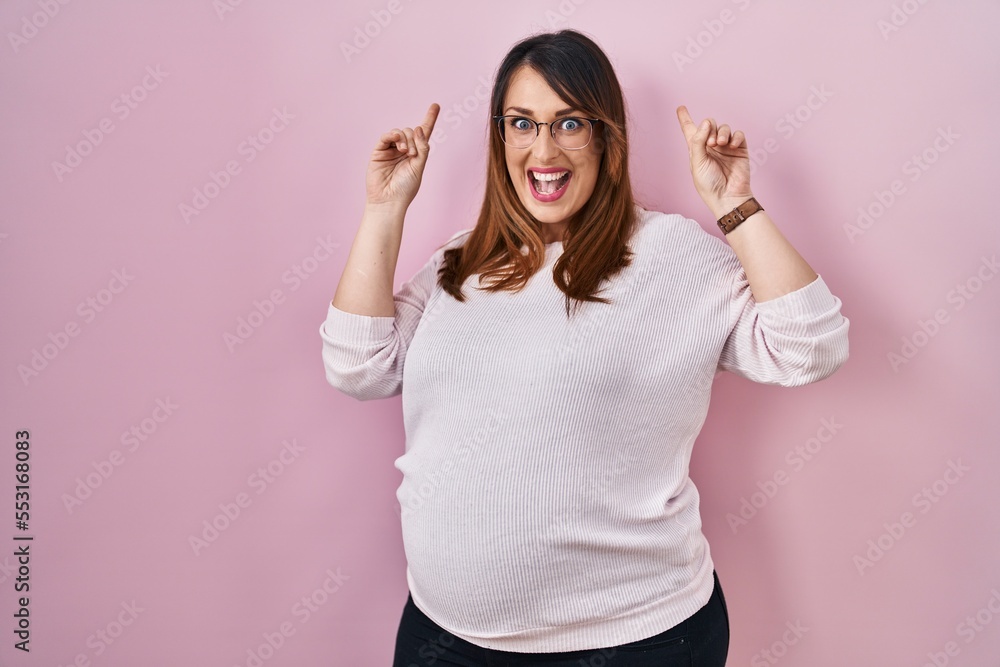 Poster pregnant woman standing over pink background smiling amazed and surprised and pointing up with finge