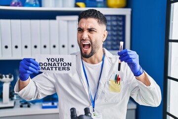 Young handsome man working at scientist laboratory holding blood sample angry and mad screaming frustrated and furious, shouting with anger. rage and aggressive concept.