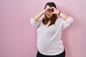 Pregnant woman standing over pink background doing heart shape with hand and fingers smiling looking through sign