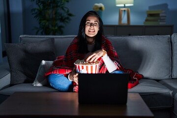 Hispanic woman eating popcorn watching a movie on the sofa winking looking at the camera with sexy...