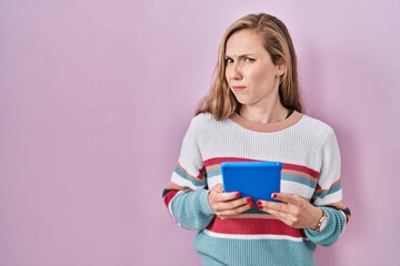 Young blonde woman holding touchpad clueless and confused expression. doubt concept.