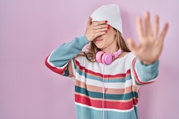 Young blonde woman standing over pink background covering eyes with hands and doing stop gesture...