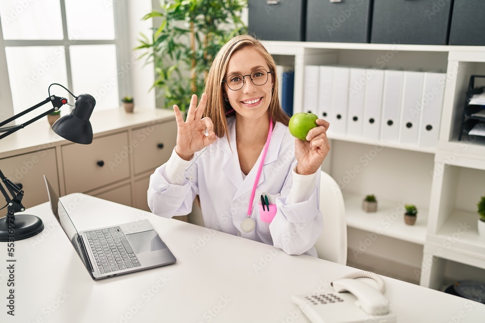 Sticker Young blonde doctor woman holding green apple at the clinic doing ok sign with fingers, smiling friendly gesturing excellent symbol