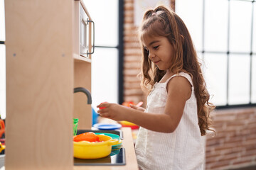 Adorable hispanic girl playing with play kitchen standing at kindergarten
